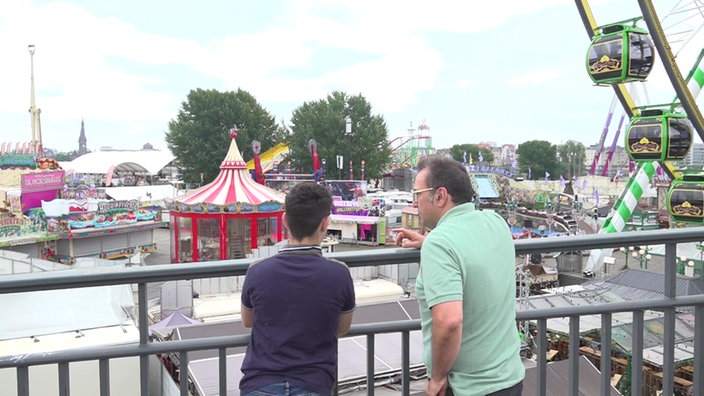Ein Mann und sein Sohn stehen an einem Geländer und schauen auf viele Fahrgeschäfte