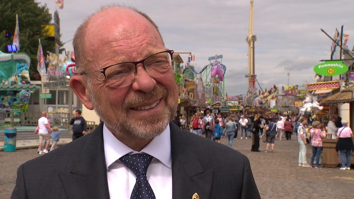Ein älterer Mann mit schütterem Haar, grauem Bart und Brille, im Anzug, vor vielen Kirmes-Fahrgeschäften