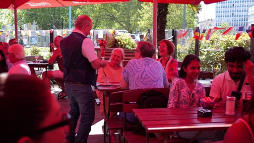 Ein Mann bringt einer Gruppe Gäste Eisbecher. Die Gäste lachen. 
