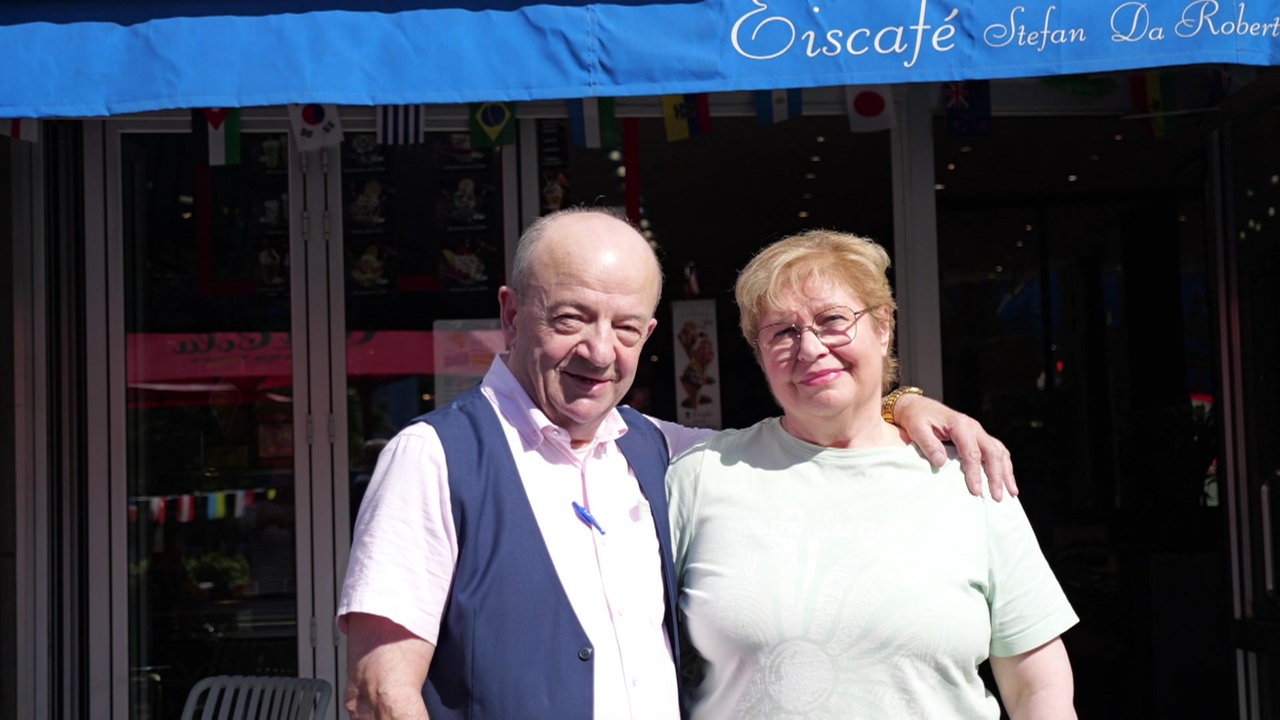 Ein Mann und eine Frau posieren vor einem Café. Die Markise liest "Eiscafé Stefan da Roberto"