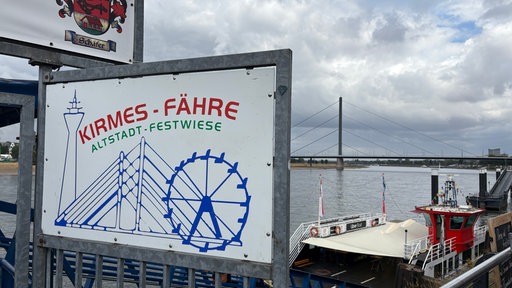 Schild mit der Aufschrift "Kirmes-Fähre - Altstadt - Festwiese" vor dem Rhein