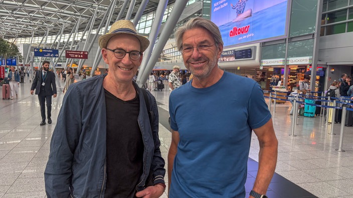 Zwei Männer stehen lächelnd in der Abflughalle des Flughafen Düsseldorf.