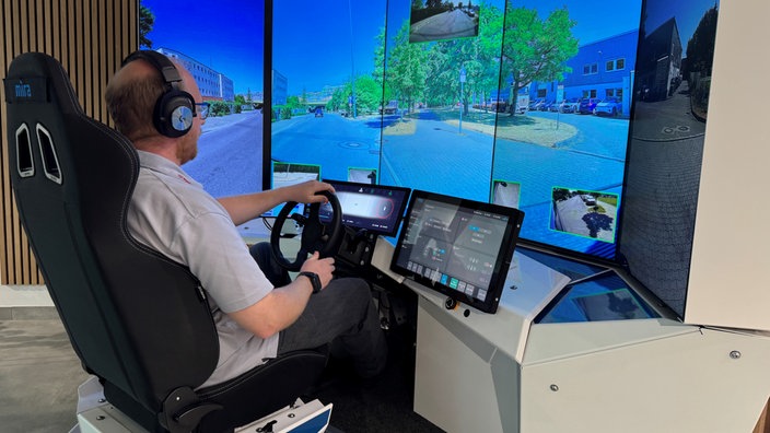 Ein Mann sitzt mit Kopfhörern vor einem großen Bildschirm und lenkt digital mit einem Lenkrad. 