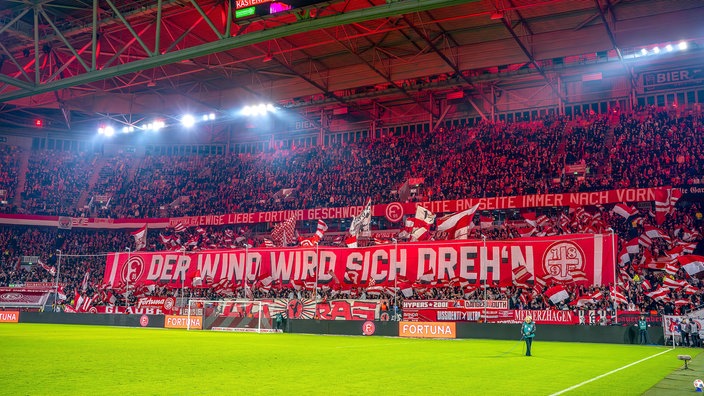 Auf dem Bild sieht man die Fankurve von Fortuna Düsseldorf mit einem großen Banner, dort steht: Der Wind wird sich drehen.