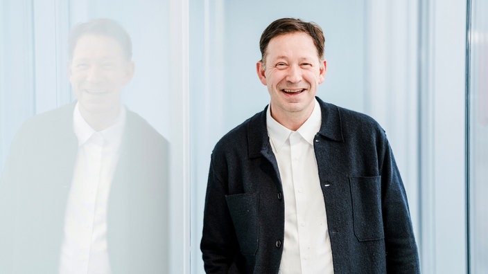 Ein lächelnder Mann mit weißem Hemd und dunkler Jacke vor einer Fensterfront