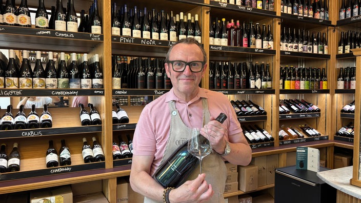 Ein Mann hält eine große Flasche und ein Weinglas in seinen Händen, im Hintergrund ein großes Weinregal