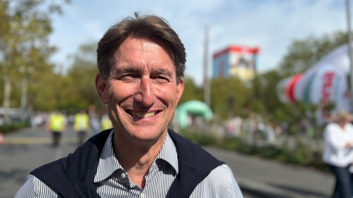 Ein Mann lächelt in die Kamera; hinter ihm sieht man unscharf eine riesige Persilflasche