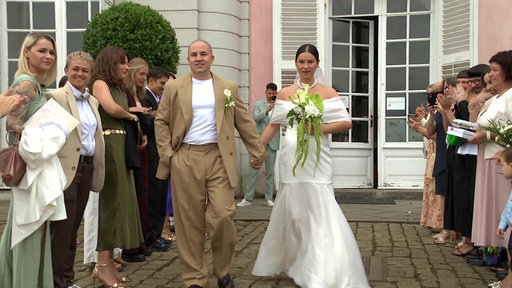 Das frischvermählte Paar vor dem Schloss Benrath. Neben ihnen sind Gäste der Hochzeit zu sehen, die Spalier stehen.