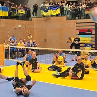 Zwei Mannschaften mit jeweils 6 Personen sitzen durch ein tiefes Volleyball- Netz getrennt. Näher an der Kamera sitzt das schwarz- tragende deutsche Team, weiter hinten die Gelb- tragenden Ukrainer. 