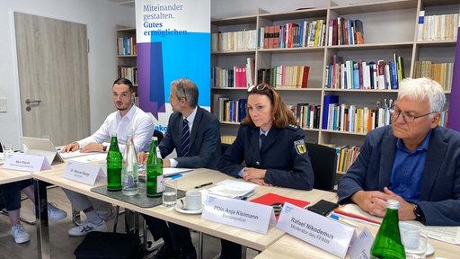 Drei Männer und eine Polizeibeamtin sitzen an einem Tisch vor einem Bücherregal, vor ihnen Namensschilder.