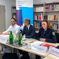 Drei Männer und eine Polizeibeamtin sitzen an einem Tisch vor einem Bücherregal, vor ihnen Namensschilder.