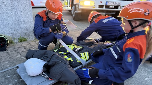 Drei Kinder der Jugendfeuerwehr führen Übungen an einer Puppe auf einer Trage durch.