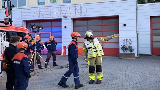 Ein Feuerwehrmann steht mit mehreren Kindern der Jugendfeuerwehr vor der Wache in Angermund und zeigt auf etwas.