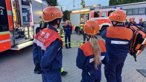 Drei Kinder der Jugendfeuerwehr stehen in Ausrüstung vor Löschfahrzeugen.