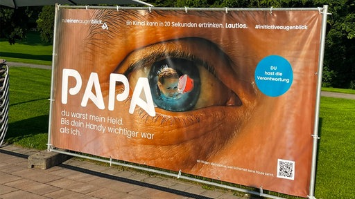Auf einem Werbeplakat ist ein Auge, darin spiegelt sich ein Kind im Wasser. Auf dem Plakat steht "Papa du warst mein Held. Bis dein Handy wichtiger war als ich" und "Ein Kind kann in 20 Sekunden ertrinken. Lautlos."
