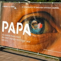 Auf einem Werbeplakat ist ein Auge, darin spiegelt sich ein Kind im Wasser. Auf dem Plakat steht "Papa du warst mein Held. Bis dein Handy wichtiger war als ich" und "Ein Kind kann in 20 Sekunden ertrinken. Lautlos."