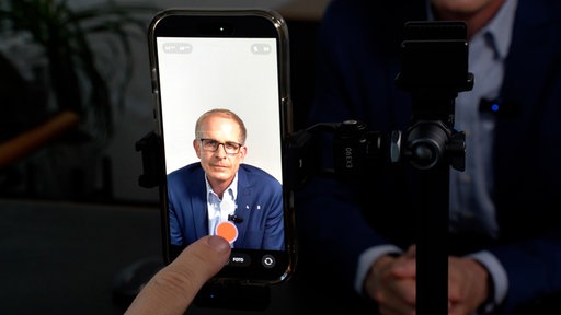 Ein Handy ist an einem Stativ befestigt, die Kamera ist geöffnet. Im Bild zu sehen ist der Langenfelder Bürgermeister Gerold Wenzens.