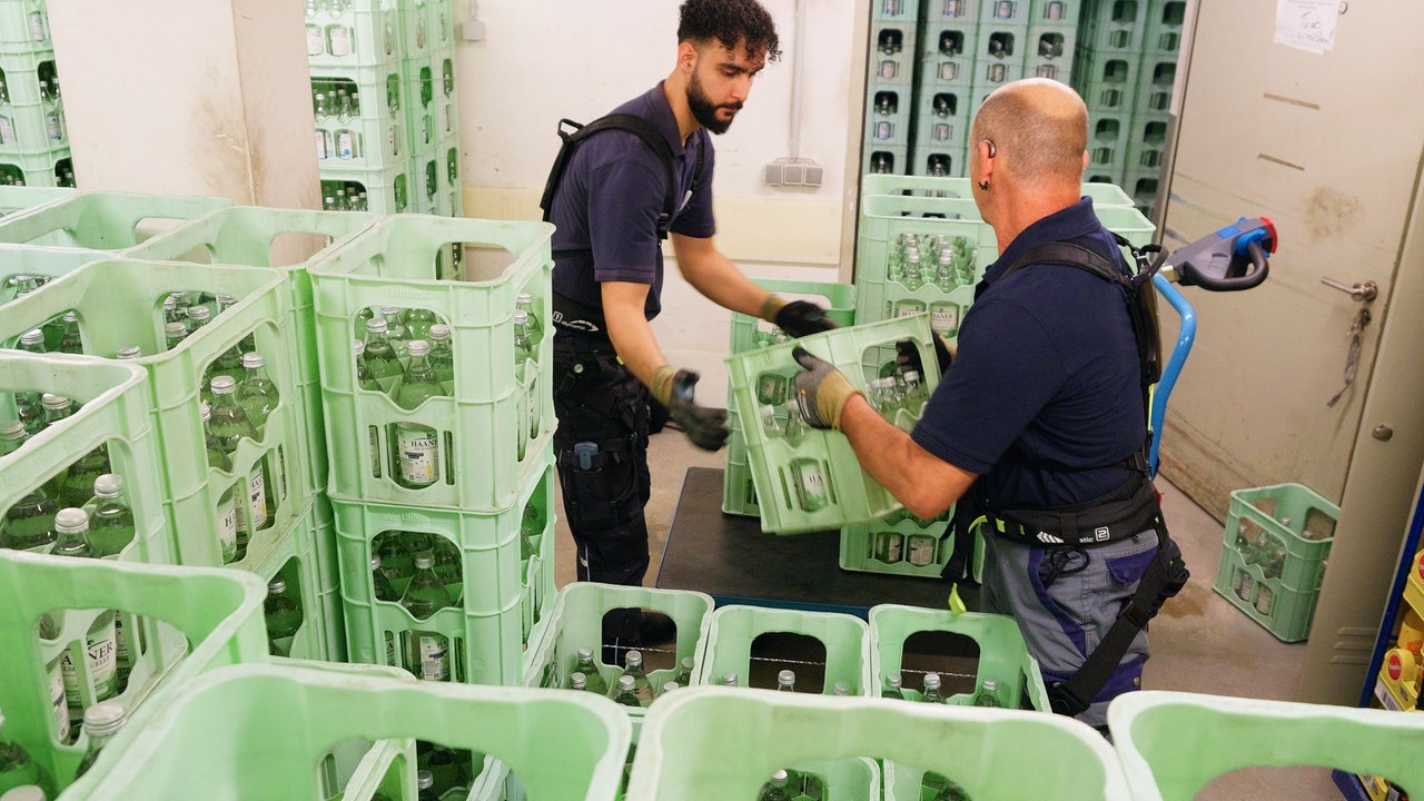 Männer transportieren Getränkekisten und tragen dabei Exoskelette auf dem Rücken. Mit Bändern und Gurten an Schultern, Rücken, Hüfte und Beinen sieht das Exoskelett aus wie ein flacher Wanderrucksack