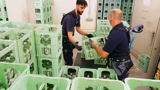 Männer transportieren Getränkekisten und tragen dabei Exoskelette auf dem Rücken. Mit Bändern und Gurten an Schultern, Rücken, Hüfte und Beinen sieht das Exoskelett aus wie ein flacher Wanderrucksack