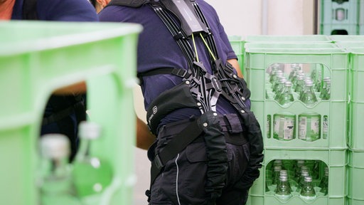 Zwischen Getränkekisten bückt sich ein Mann. Im Ausschnitt ist nur sein Rücken mit dem Exoskelett zu sehen. Die gelben Bänder am Exoskeltt verbinden die Rückenplatte und den Hüftgurt
