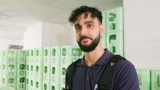 Mann mit schwarzen Haaren steht vor Getränkekästen und lächelt richtung Kamera.