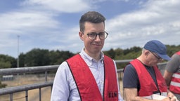 Auf dem Bild sieht man einen Mann mit roter Warnweste, der zuversichtlich in die Kamera schaut, im Hintergrund sieht man eine Baustelle.