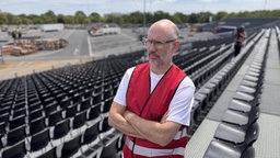 Auf dem Bild sieht man einen Mann mit Brille in einer roten Warnweste, der auf einer Sitzplatz-Tribüne steht und in die Ferne blickt.
