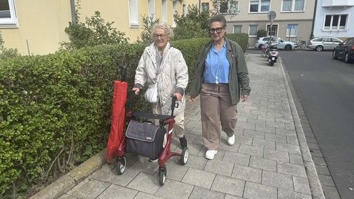 Zwei Frauen laufen über den Bürgersteig, die ältere von beiden schiebt einen Rollator.