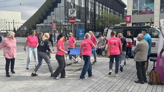 Ein Dutzend Frauen stehen auf einem Platz um eine Tischtennisplatte herum. Die Frauen tragen pinke T-Shirts.