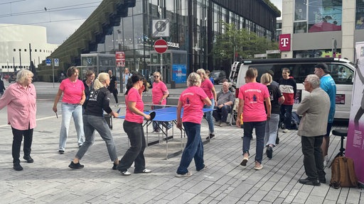Ein Dutzend Frauen stehen auf einem Platz um eine Tischtennisplatte herum. Die Frauen tragen pinke T-Shirts.