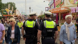 Zwei Polizisten von Hinten zu Sehen. Sie laufen über einen gut beschäftigten Kirmesgrund.