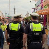Zwei Polizisten von Hinten zu Sehen. Sie laufen über einen gut beschäftigten Kirmesgrund.