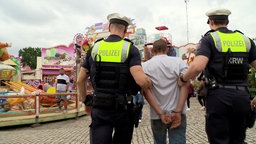Zwei Polizisten führen eine Person in grauem T Shirt und Handschellen ab.