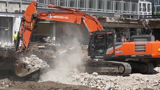 Ein Bagger räumt Trümmer eines Abrisses beiseite. Er ist orange und steht auf einer Baustelle.