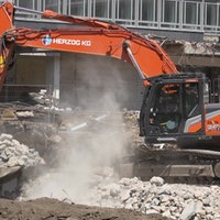 Ein Bagger räumt Trümmer eines Abrisses beiseite. Er ist orange und steht auf einer Baustelle.