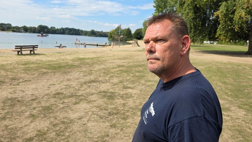 Frank Bauer, Leiter der Strandbäder am Unterbacher See in Düsseldorf, ist im Portät zu sehen. Im Hintergrund liegt der Unterbacher See