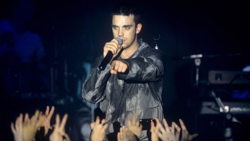 Der Musiker Robbie Williams steht auf der Bühne und singt in ein Mikrofon. Vor ihm recken sich die Hände von Zuschauern in die Luft.