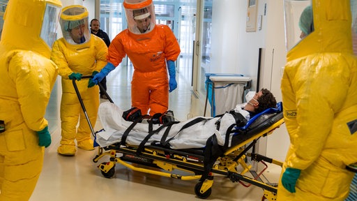 Mehrere Menschen in Ganzkörperschutzanzügen schieben ein Mensch auf einer Trage in einen Raum