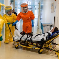 Mehrere Menschen in Ganzkörperschutzanzügen schieben ein Mensch auf einer Trage in einen Raum