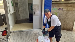 Shorty steht vor aufgestellten mobilen Duschen mit Putzmitteln in der Hand.