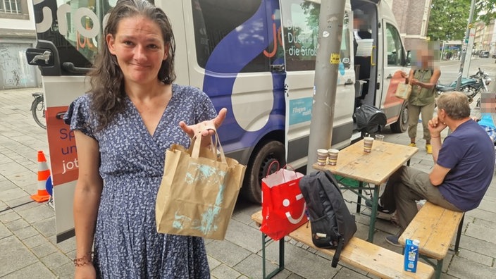 Eine Frau steht vor einem Transporter und hält eine Papiertüte vor sich.