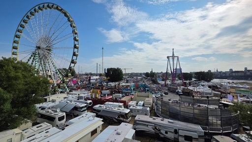 Riesenrad, viele Fahrgeschäfte und Campingwagen stehen auf der Wiese am Rhein.