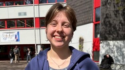 Eine junge Frau in T-Shirt und blauer Strickjacke steht vor der Mensa und lächelt in die Kamera
