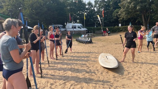Eine Gruppe steht vor einer Frau und hört ihr zu. Zu ihren Füßen liegen SUP-Boards.