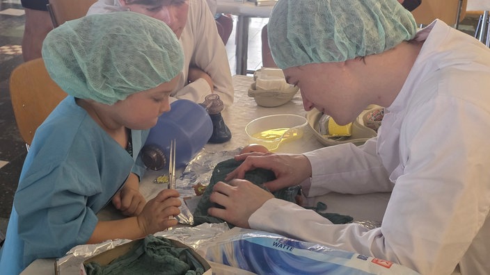 Ein Mädchen und ein Student beugen sich in OP-Klamotten über ein Kuscheltier auf einem Tisch
