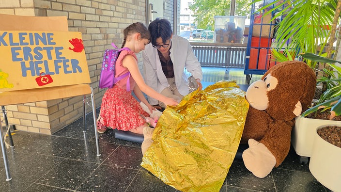 EIn Mädchen und ein Mann in weißem Kittel kniend vor einem großen Teddybär, auf dem eine Rettungsdecke