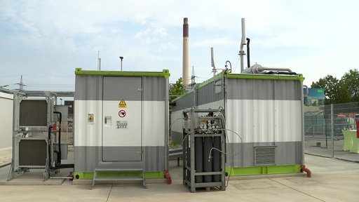 Vor zwei weiß-grauen Containern mit grünem Dach steht ein schwarzer Kasten mit vielen Kabeln.