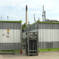 Vor zwei weiß-grauen Containern mit grünem Dach steht ein schwarzer Kasten mit vielen Kabeln.