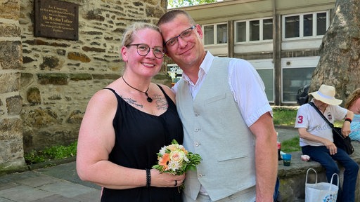 Ein Paar steht mit Strauß und festlicher Kleidung vor einer Kirche.