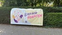 Ein Banner mit der Aufschrift "Einfach Heiraten" ist an einer grünen Hecke befestigt.